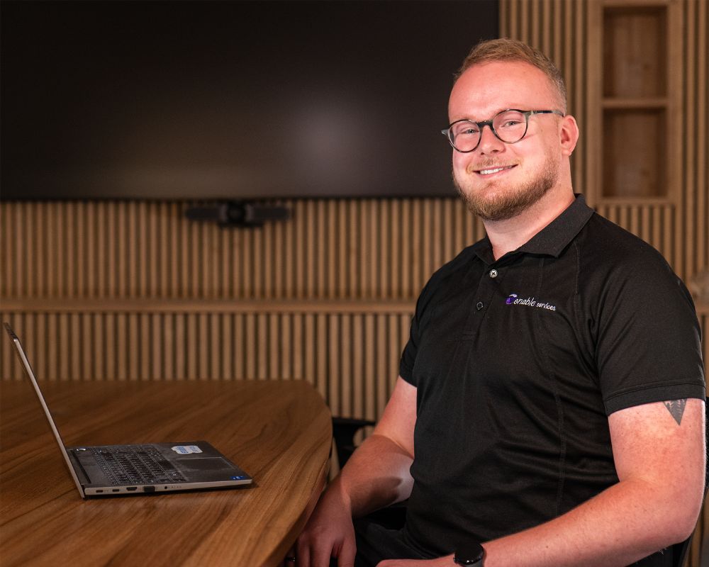 Man sitting and smiling at camera in office with laptop on desk infront of him