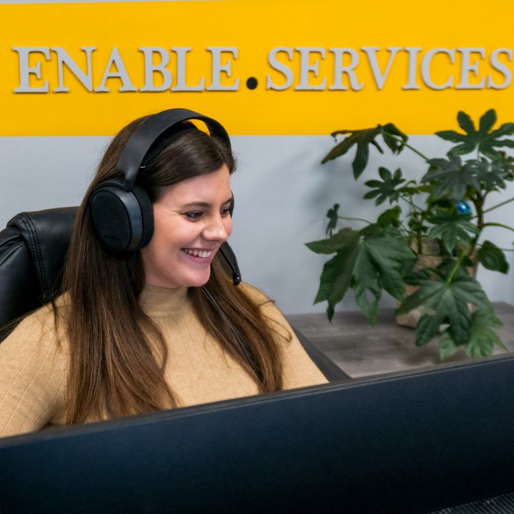 Woman in office wearing headset and smiling at desk in front of enable.services wall sign